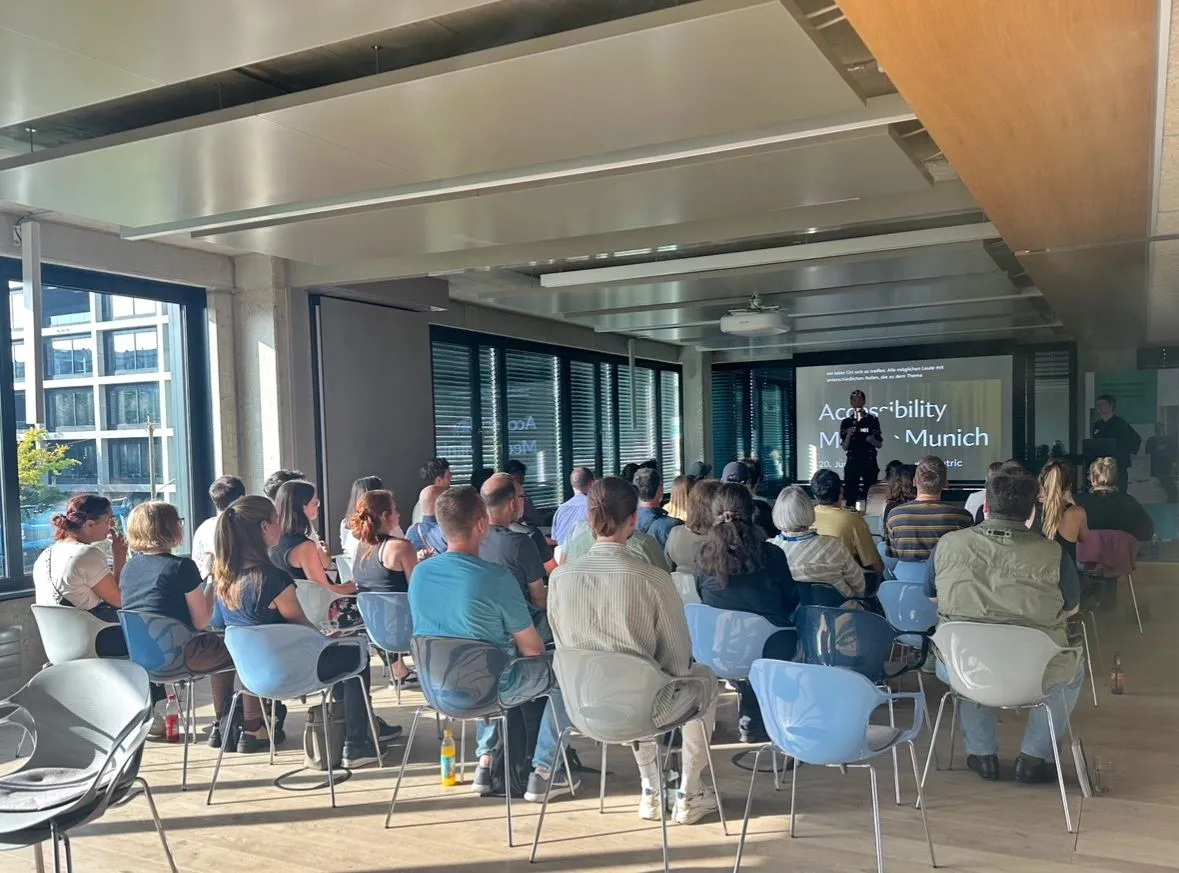 In a sunlit conference room filled with around forty attendees, a screen at the far end  displays a slide titled 'Accessibility Meetup Munich'. The silhouettes of two presenters are visible on a small stage beneath the screen.