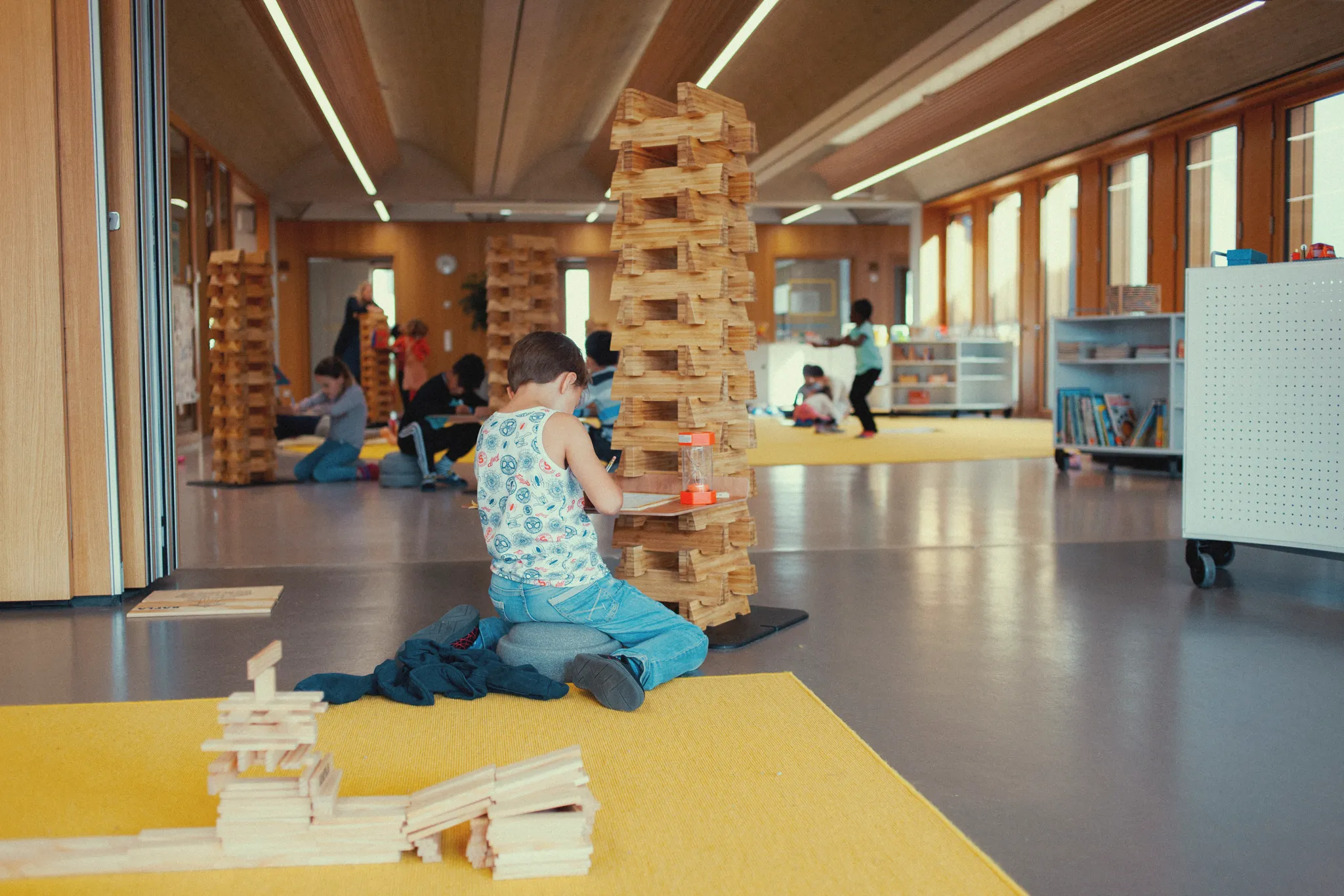 Ein Schüler sitzt in einem großen offenen Raum und schreibt. Hinter ihm auf einem großen gelben Teppich ein Turm aus Holzbausteinen, mit denen er vieleicht gerade noch gespielt hat. In der Tiefe des Raumes weitere SchülerInnen die sich konzentriert beschäftigen.